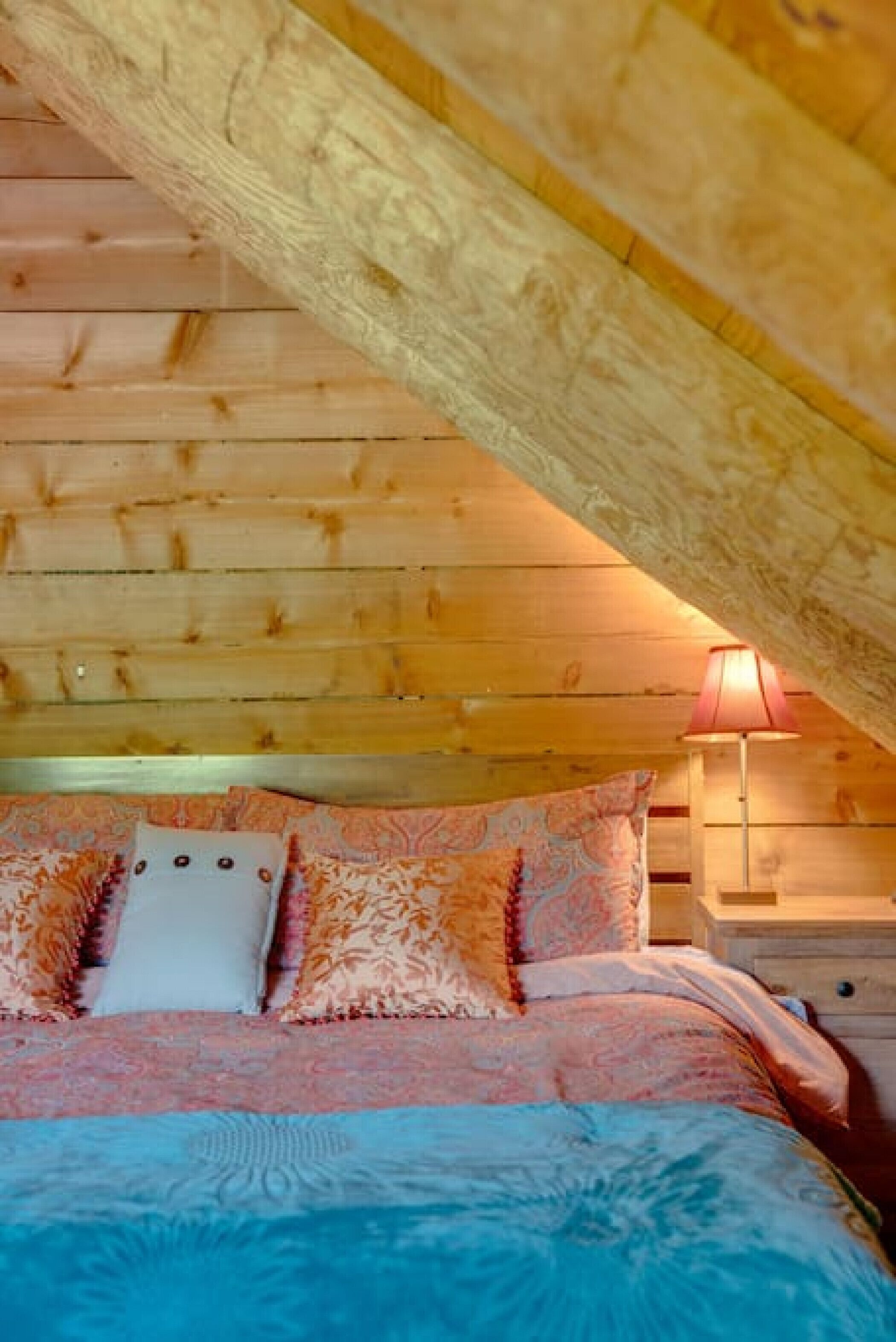 A cozy bedroom with a wooden ceiling, a bed with patterned bedding, and a bedside lamp.