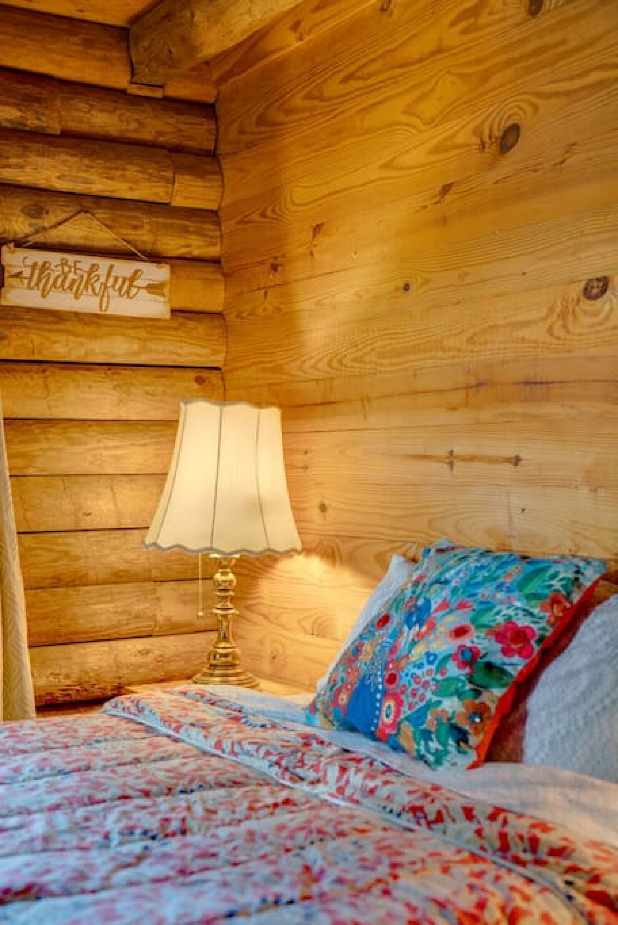 A wooden cabin interior with a bed, a lamp, and a 'thankful' sign on the wall.