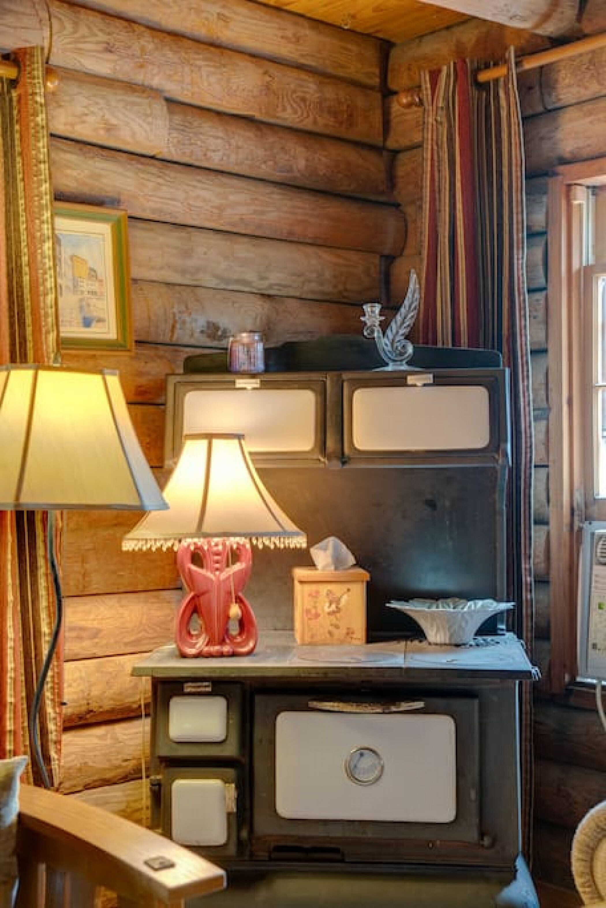 A rustic cabin interior with a wooden wall, a vintage stove, a lamp, and a framed picture.