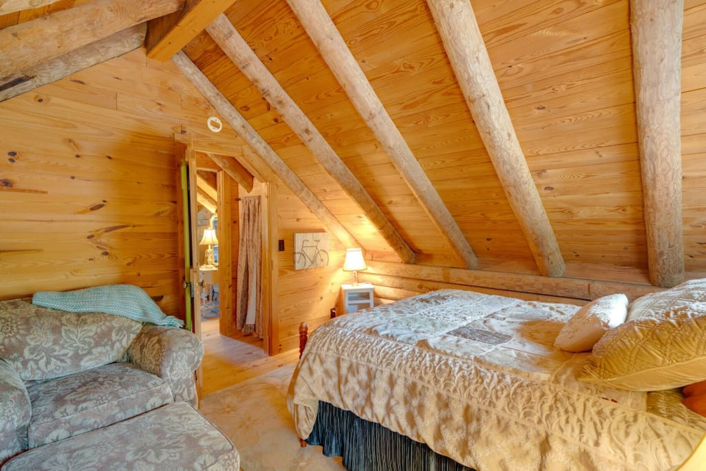 Un dormitorio rústico tipo cabaña con techo de madera, cama con colcha estampada, un sofá y una bicicleta al fondo.
