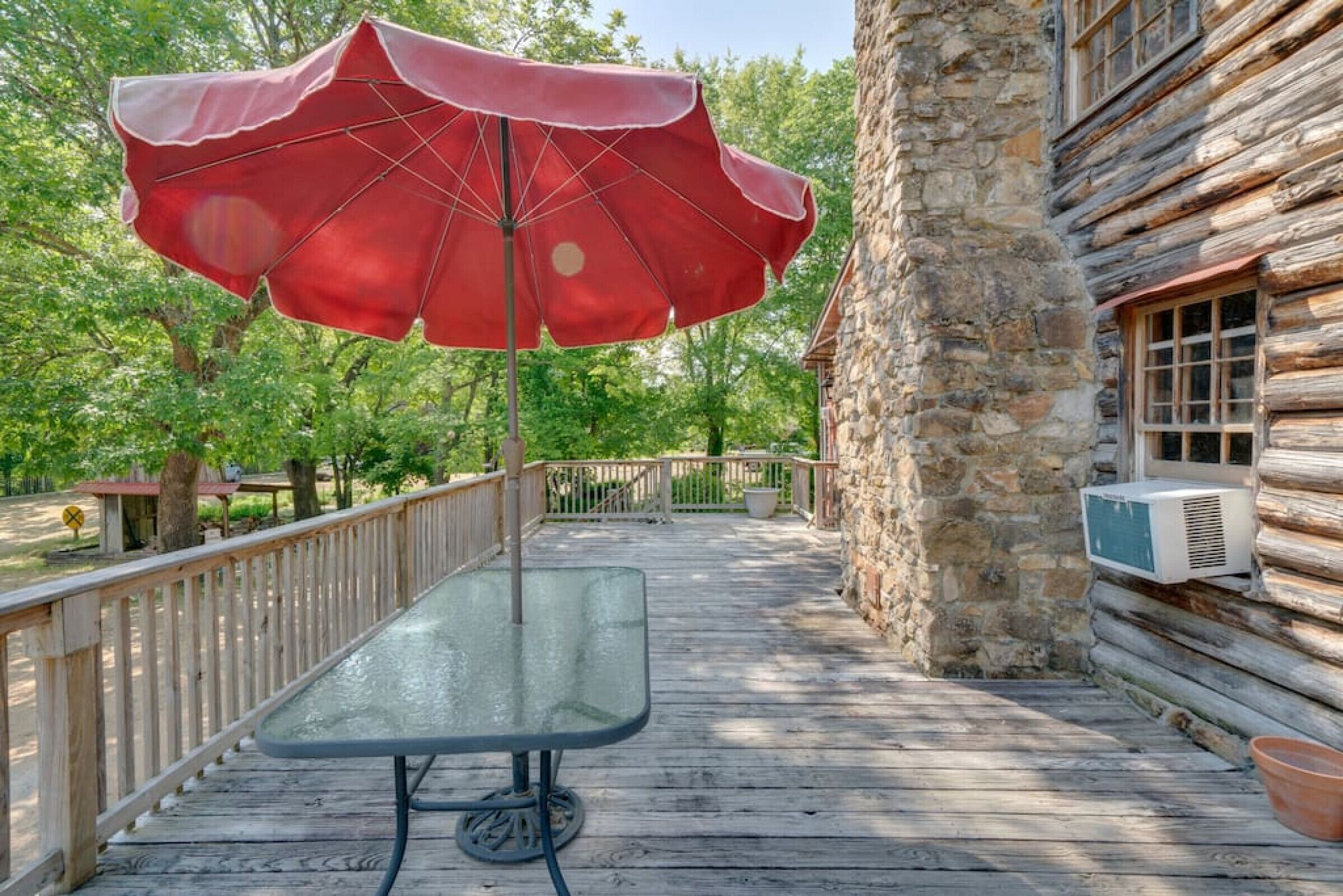 A wooden deck with a table and a red umbrella, surrounded by stone walls and greenery.