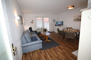 A modern living room with a grey sofa, wooden flooring, a dining table with chairs, a TV, and a window with blinds.