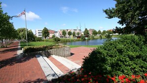 A paved walkway with a stone wall and flower beds, leading to a body of water with buildings in the background. - Beautiful, bright vacation apartment with swimming pool, Residence Kormoran15 (Cuxhaven)