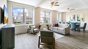 A spacious living room with a large window, a ceiling fan, and a dining area with blue chairs. - Spacious Condo in California Building (New Orleans)