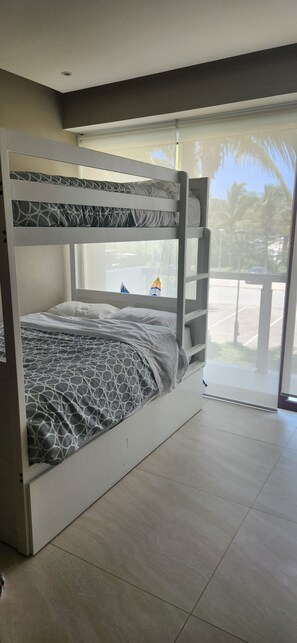 A bunk bed with a patterned bedspread, a window with a view of palm trees, and a tiled floor.