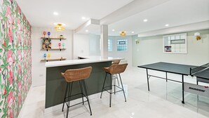 Una cocina moderna con barra, dos taburetes de mimbre, una mesa de ping-pong y una pared decorativa con estantes que exhiben diversos objetos.