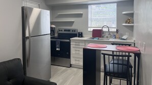 Een compacte keuken met een koelkast, fornuis, magnetron en spoelbak. Er is een raam met jaloezieën, een klein aanrecht en een stoel.