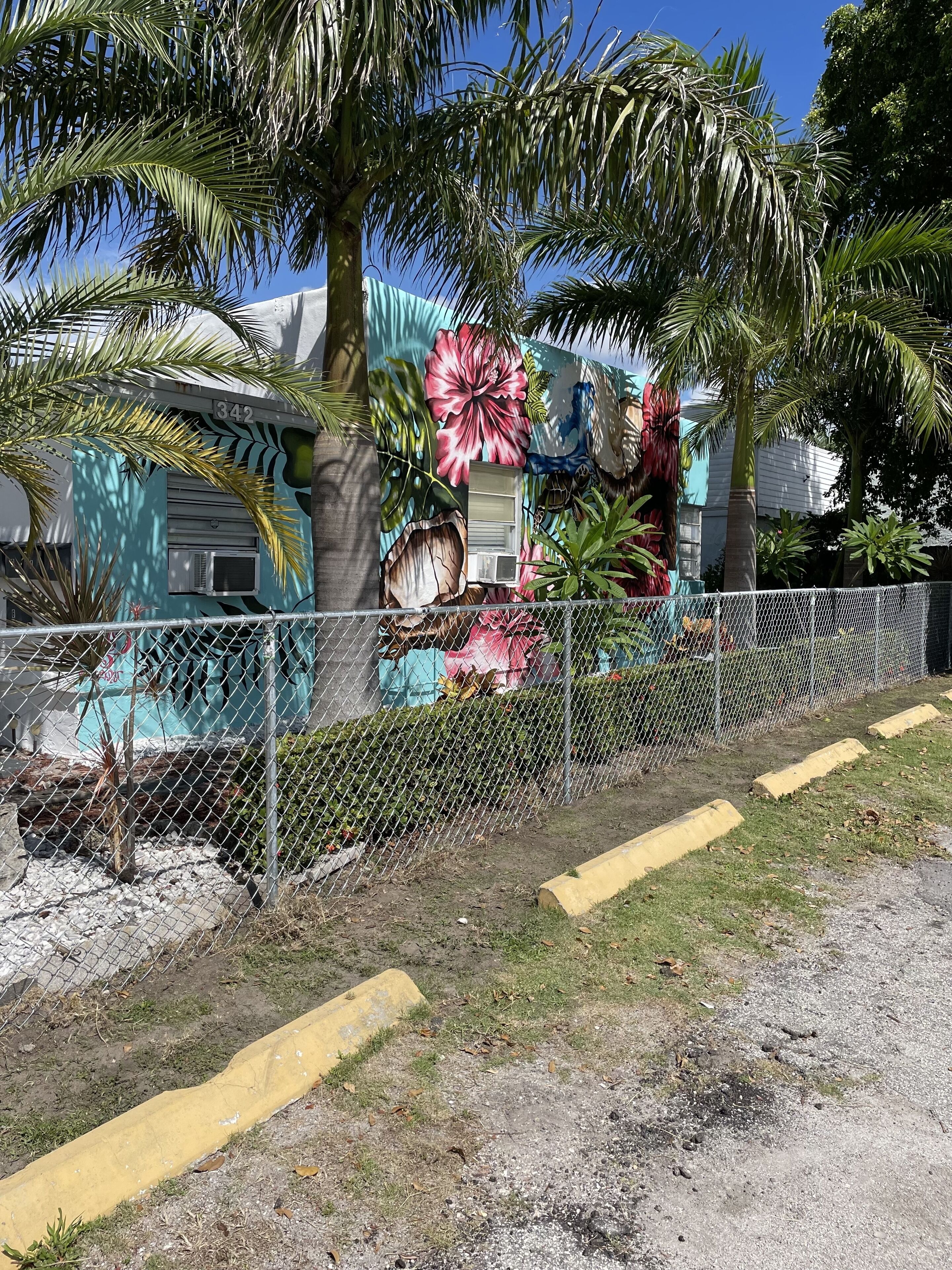 Un espace clos, agrémenté d’une fresque représentant des fleurs tropicales et des palmiers, un bâtiment à la façade turquoise et un ciel d’un bleu limpide.
