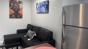 Een moderne woonkamer met een zwarte bank, een bijzettafeltje en een roestvrijstalen koelkast. Er hangen twee ingelijste kunstwerken aan de muur.