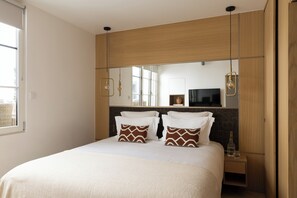 A modern hotel room with a large bed, wooden headboard, and a large mirror. There is a TV on the wall and a small table with a bottle and glass. - HIGHSTAY - Luxury Serviced Apartments - Saint-Honoré (Paris)