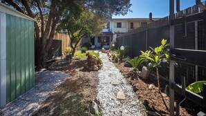 Un étroit sentier de gravier mène à une maison dotée d’une cour clôturée, entourée d’arbres et de plantes.