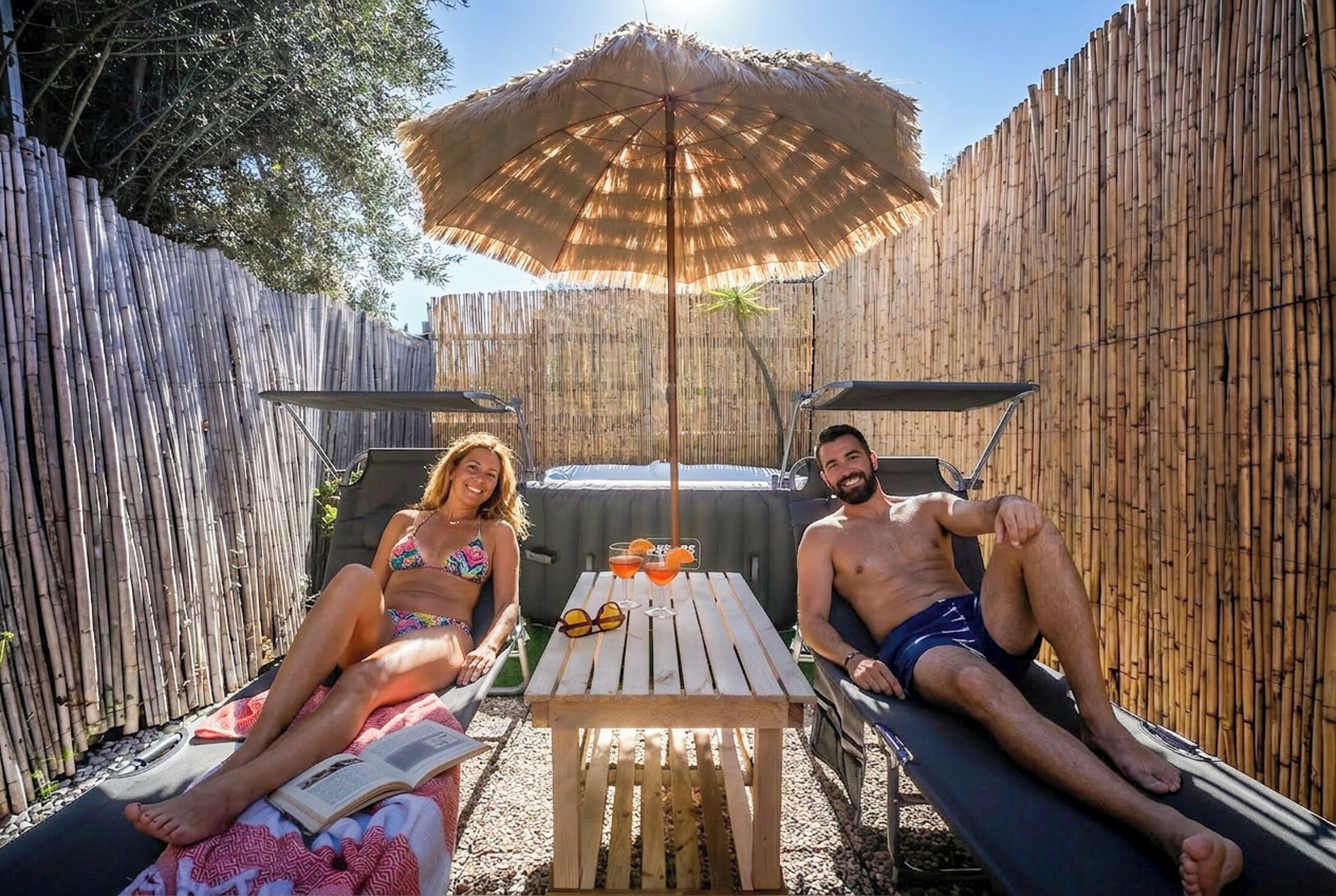 Deux personnes se détendent sur des transats, à l’ombre d’une clôture en bambou, une table placée entre elles servant de support à des boissons et des lunettes de soleil.