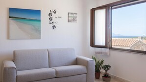 A living room with a beige sofa, a window with a view of the sea, and two potted plants.
