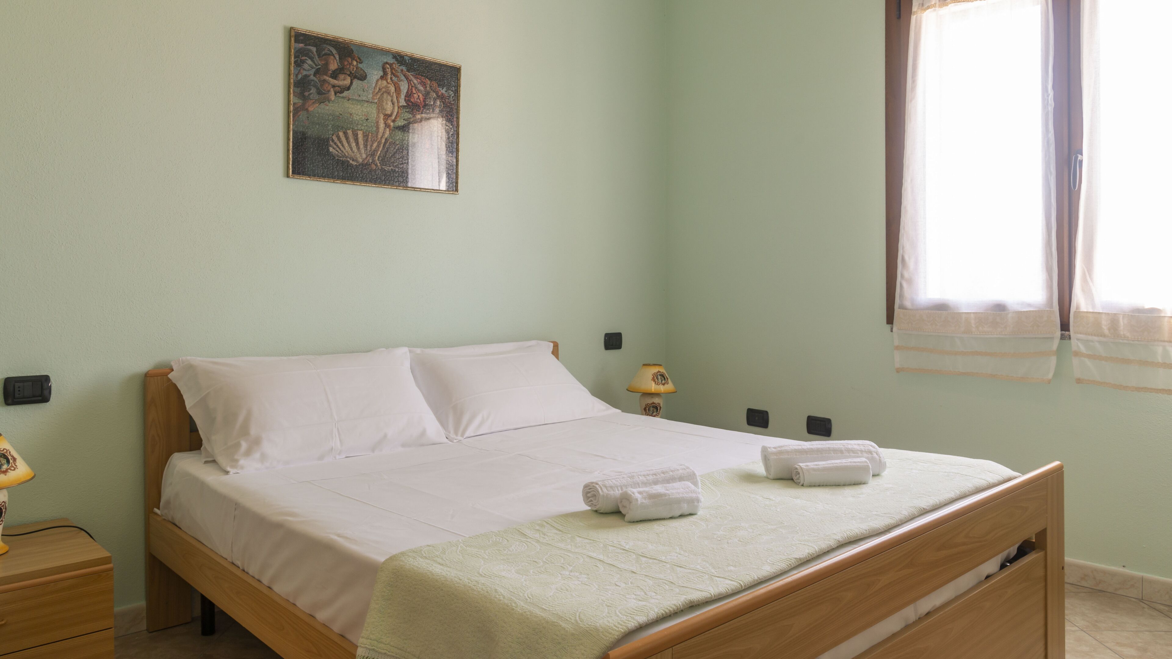 Een slaapkamer met een bed, nachtkastje, lamp, schilderij aan de muur en raam met gordijnen.