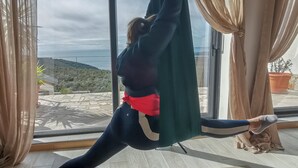 Eine Person übt Aerial Yoga in einem Raum mit großen Fenstern, Holzboden und Vorhängen.