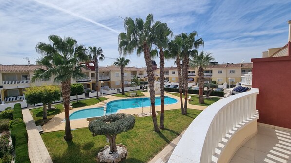 A pool area with palm trees, a well-maintained lawn, and a building with balconies in the background.