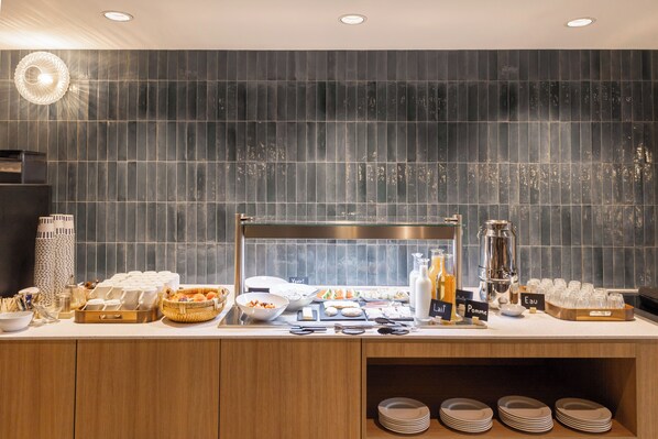Una mesa de buffet con distintos alimentos, utensilios, una cafetera y una vitrina de vidrio.