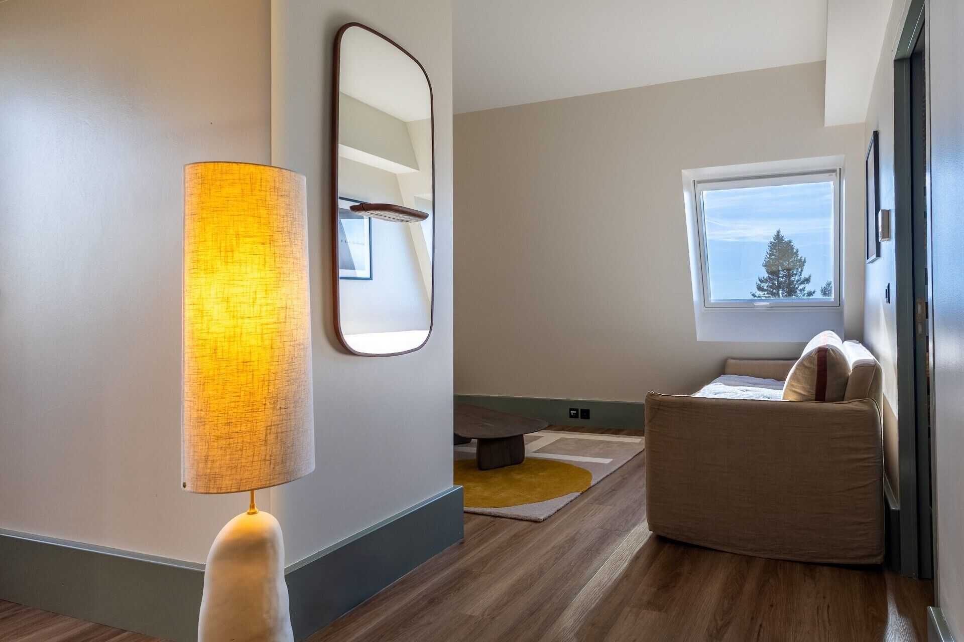 A modern living room with a sofa, a lamp, a mirror, and a window with a view of trees.