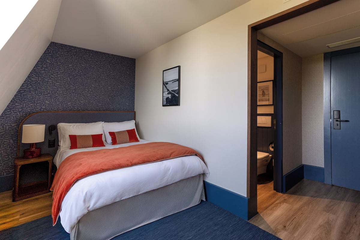 Une chambre à coucher comprenant un lit, une table de chevet, une lampe fixée au mur et une porte donnant accès à une salle de bain.