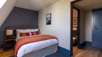 Une chambre à coucher comprenant un lit, une table de chevet, une lampe fixée au mur et une porte donnant accès à une salle de bain.
