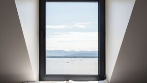 Un dormitorio con un ventanal grande, una cama con ropa de cama blanca y vistas a un cuerpo de agua y montañas.