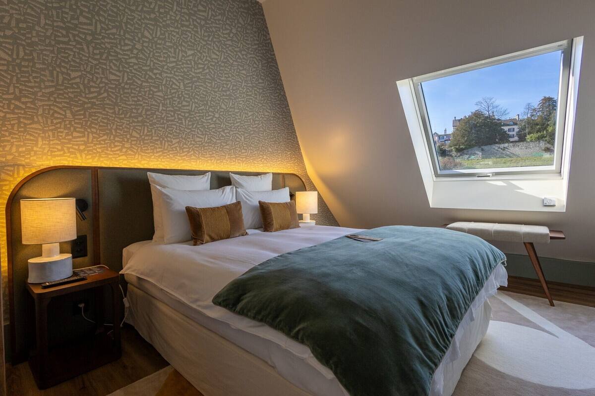 Une chambre avec un plafond en pente, une grande fenêtre, un lit recouvert de draps blancs et deux lampes de chevet.