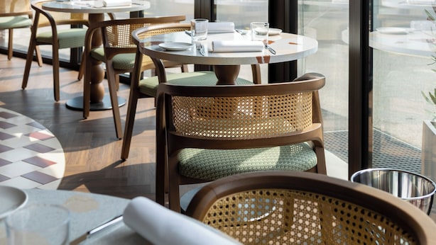 Un restaurante con mesas redondas y sillas de mimbre, amplias ventanas con vistas al mar y a las montañas.