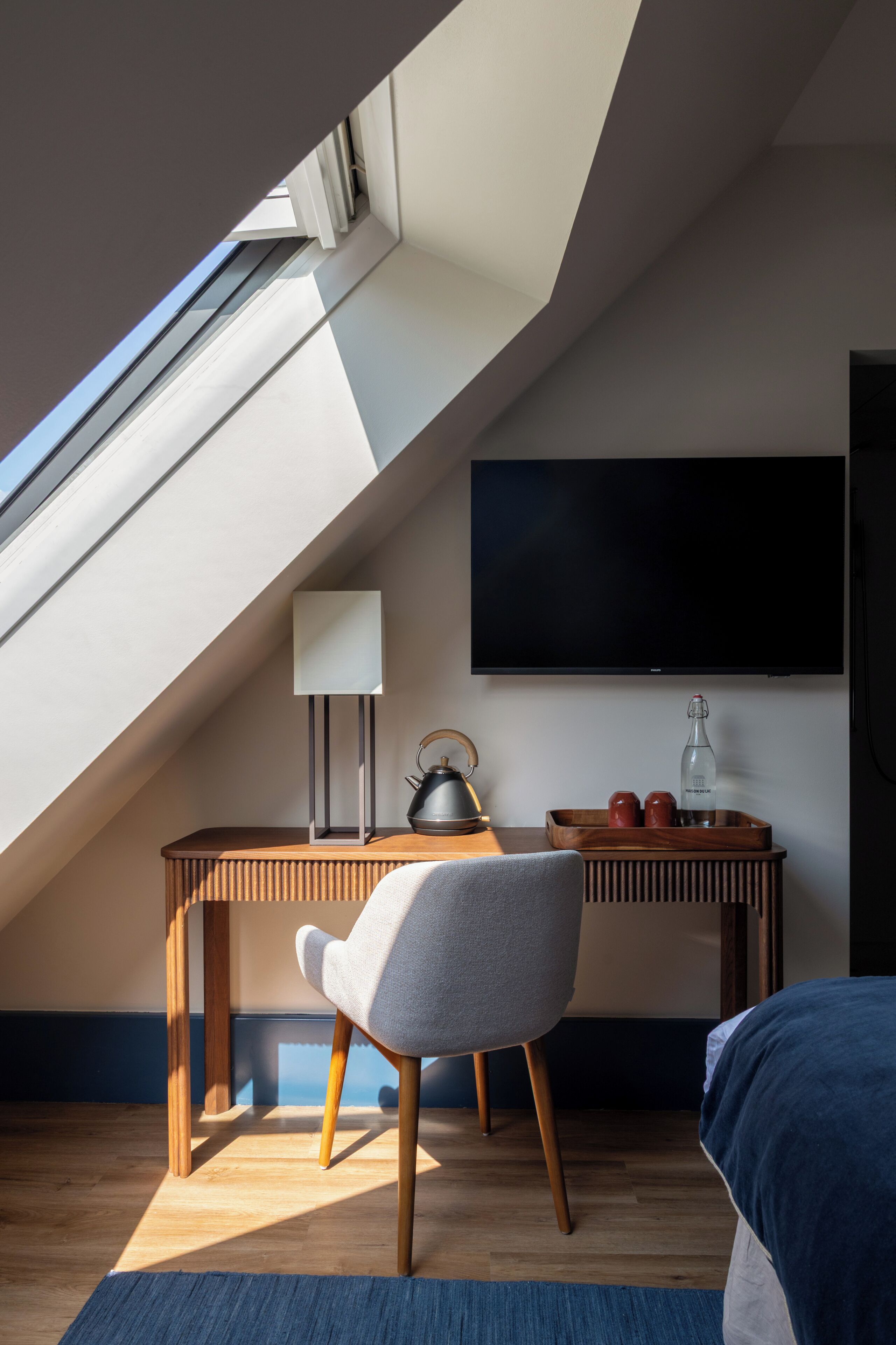 Een moderne kamer met een schuine kap, een flatscreen televisie aan de muur, een houten bureau met lamp, een stoel en een bed met een blauwe deken.
