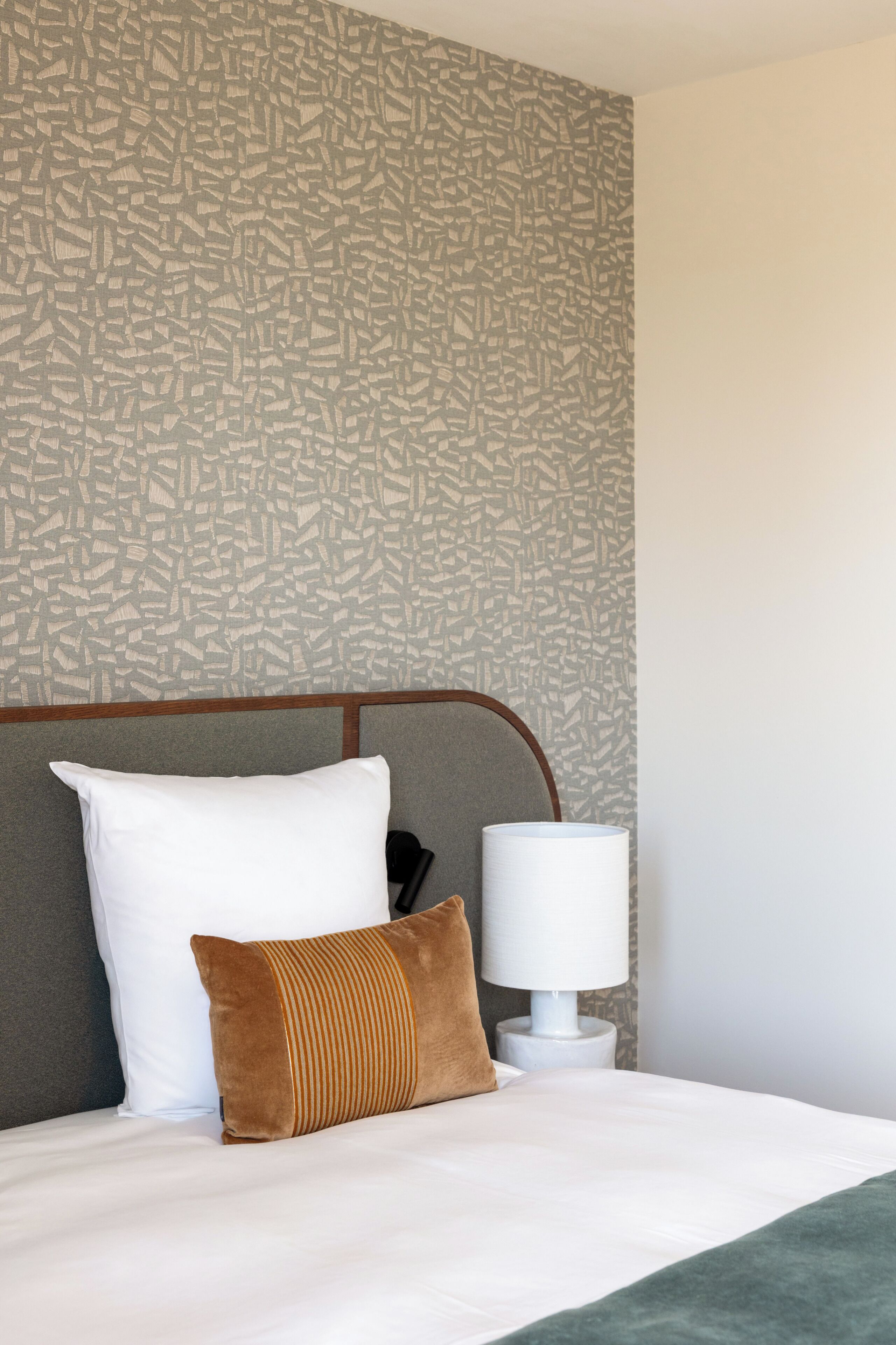 Una cama bien tendida con almohadas blancas y marrones, una lámpara blanca sobre una mesita de noche y una pared con diseño.