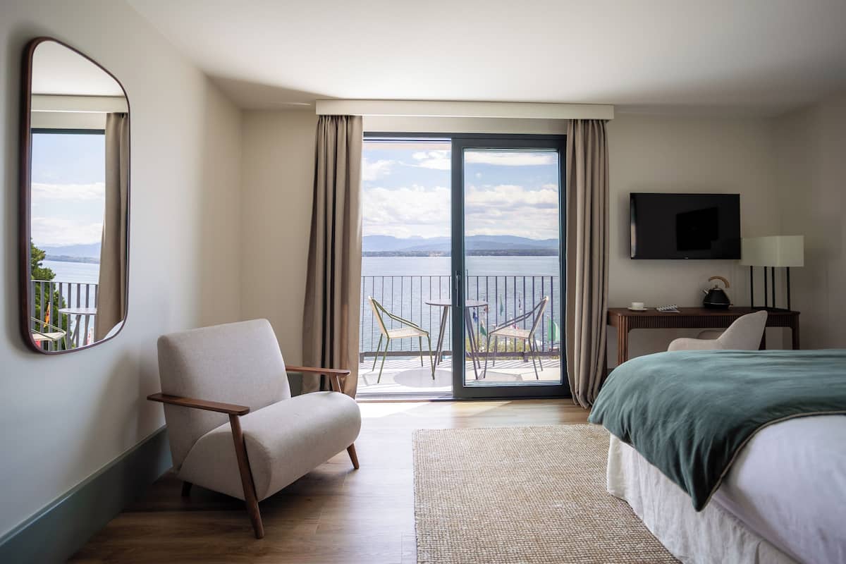 Une chambre d’hôtel avec un lit, une chaise, un bureau avec une lampe, une télévision et un balcon donnant sur la mer.