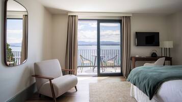 A hotel room with a bed, a chair, a desk with a lamp, a TV, and a balcony with a view of the sea.