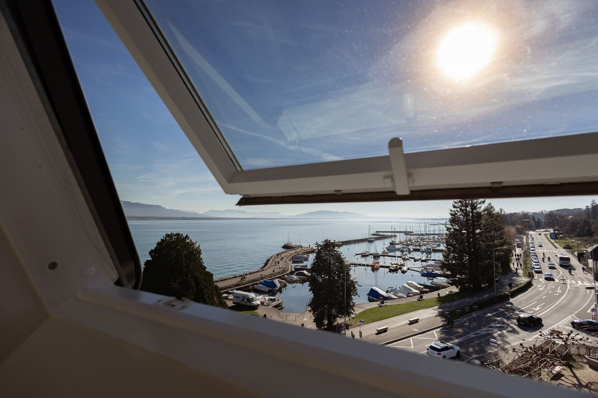 Blick durch ein Fenster auf einen Yachthafen mit vertäuten Booten, eine Straße mit Fahrzeugen und einem klaren Himmel.