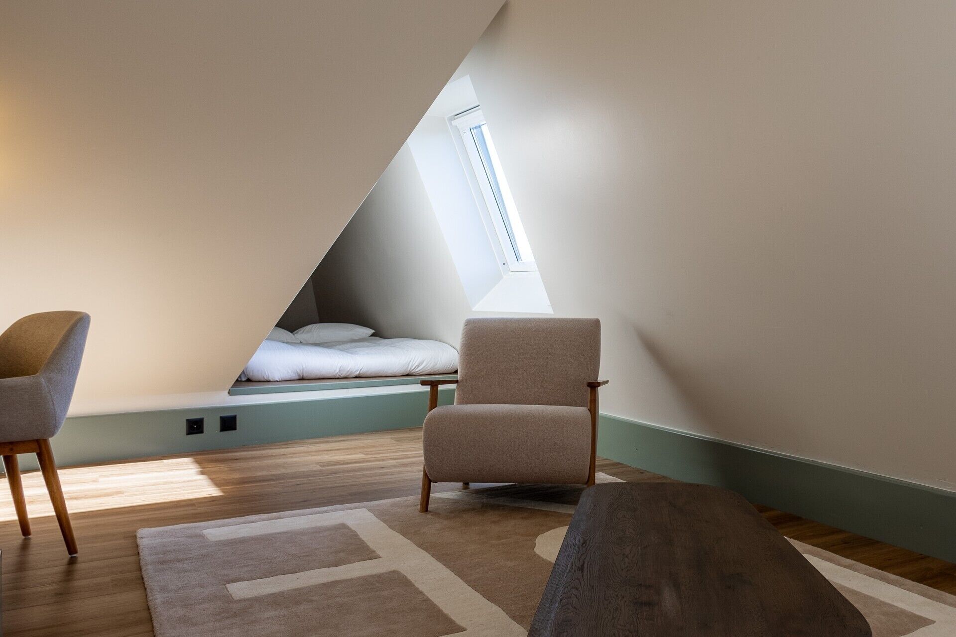 Habitación pequeña con techo inclinado, una cama, dos sillas y un ventanal grande.