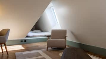 Habitación pequeña con techo inclinado, una cama, dos sillas y un ventanal grande.