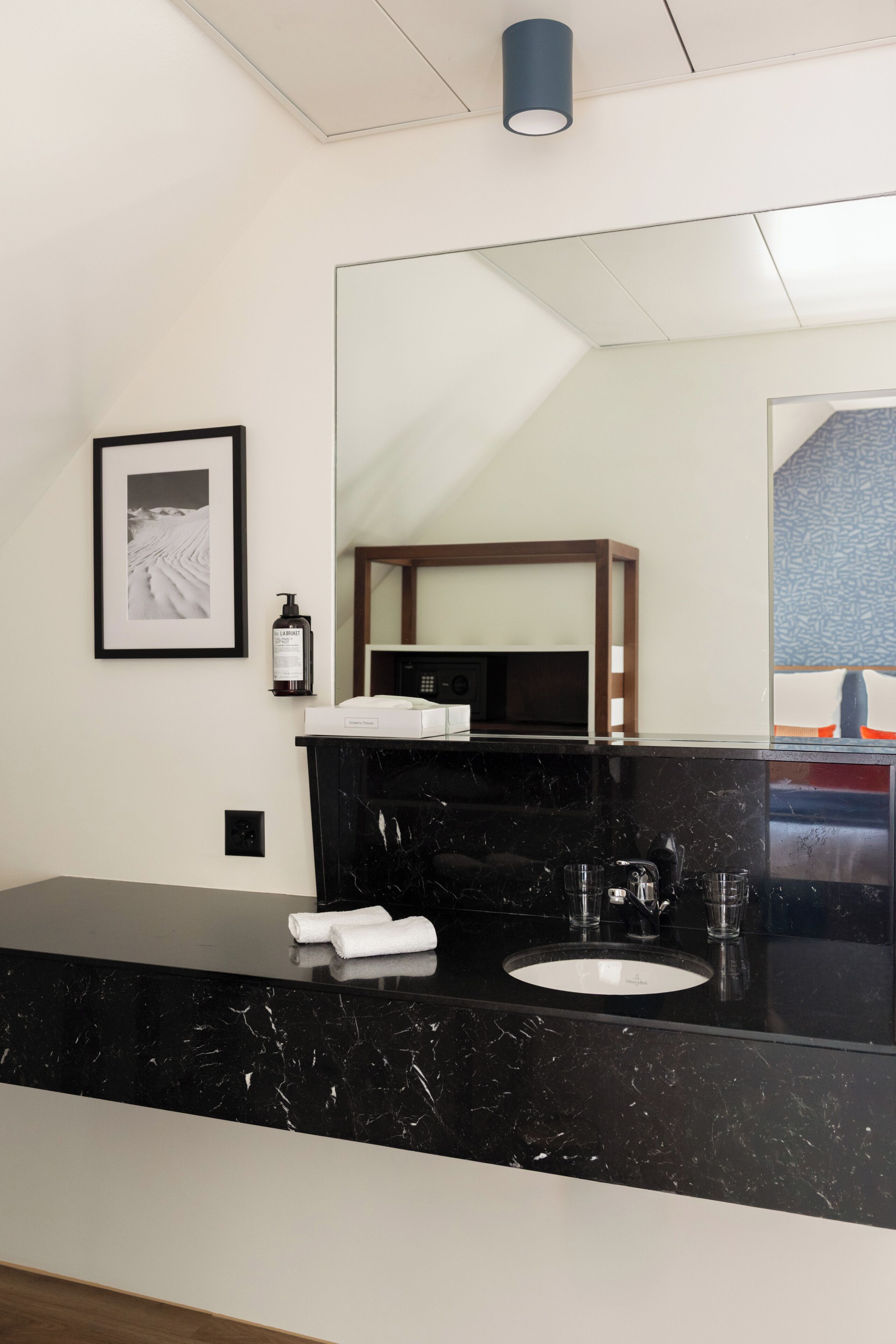A modern bathroom with a black marble countertop, a white sink, and a large mirror. There is a framed picture on the wall and a bottle of hand sanitizer.