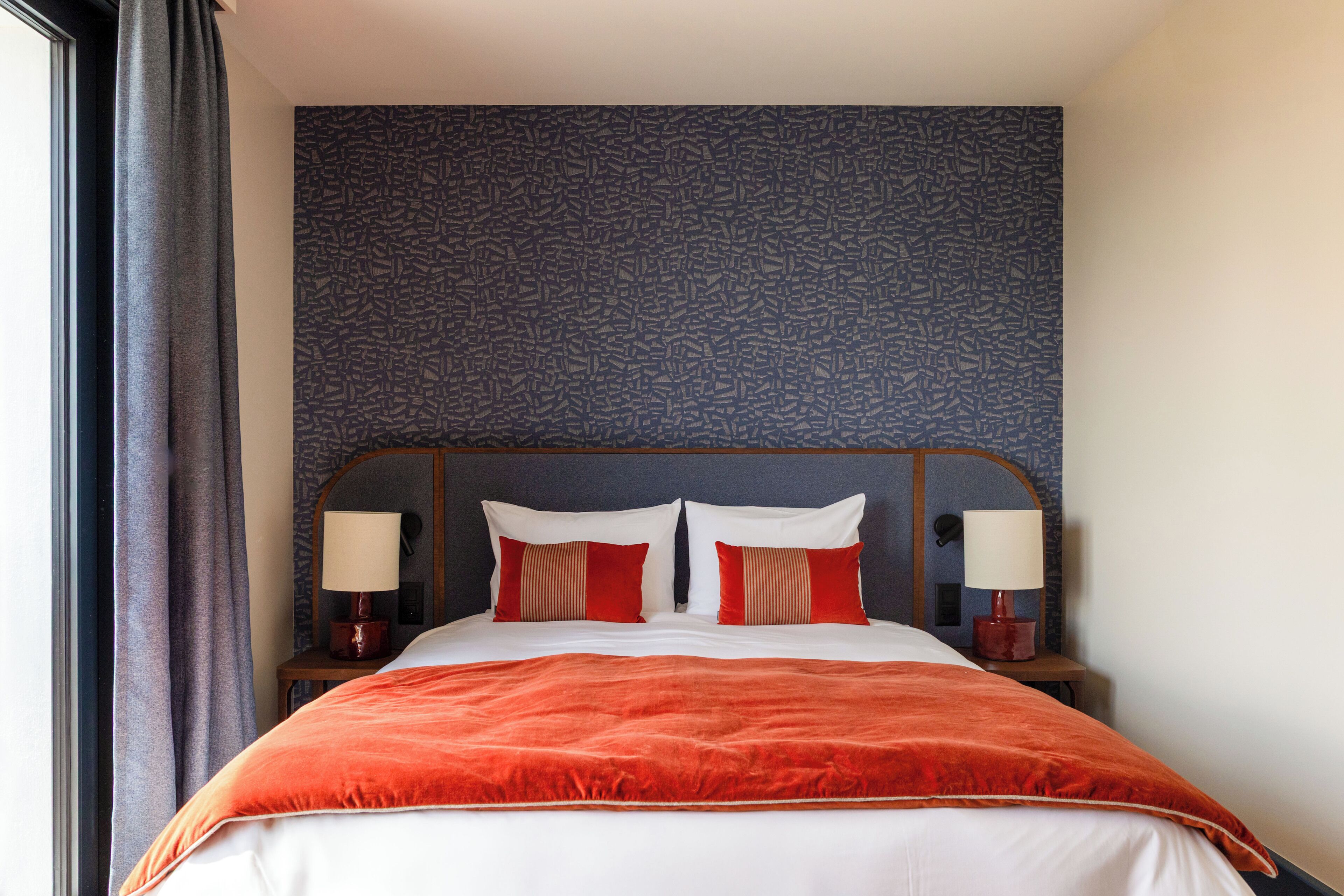 Una habitación de hotel con una cama grande, dos mesitas de noche con lámparas, un papel tapiz decorativo y una ventana con cortinas.