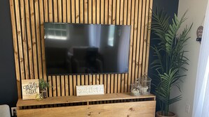 Ein Wohnzimmer mit einer Wand aus Holzleisten, einem Flachbildfernseher, einem Glastisch mit Büchern und einer Topfpflanze.
