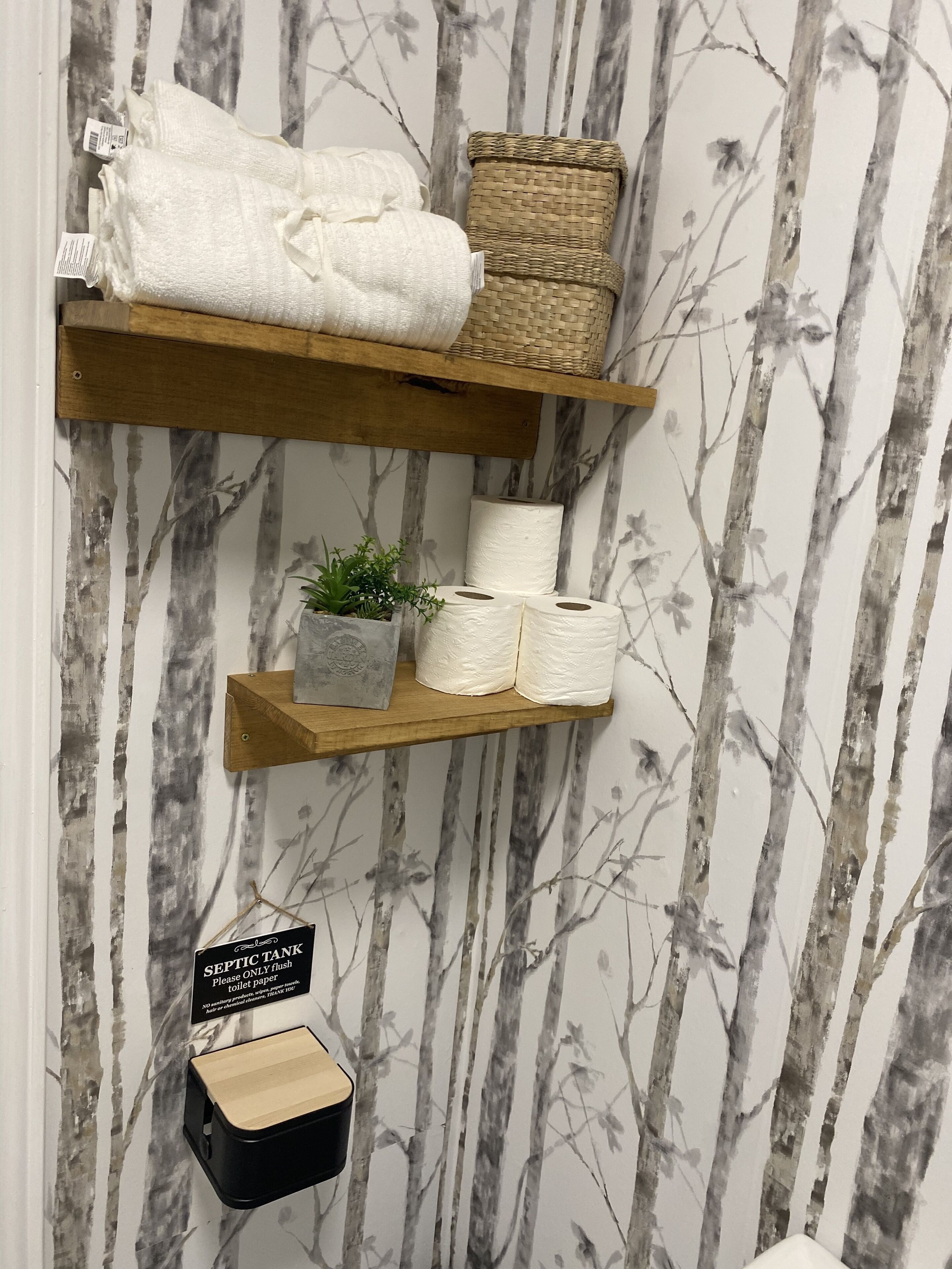 Zwei Holzregale mit Handtüchern, Toilettenpapier, einer kleinen Pflanze und einem Schild, das eine Trockentoilette kennzeichnet.