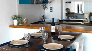 A modern kitchen with a dining area. It includes a counter with a bottle of wine, glasses, and plates. There is a blue cabinet above the counter and a hanging light fixture.