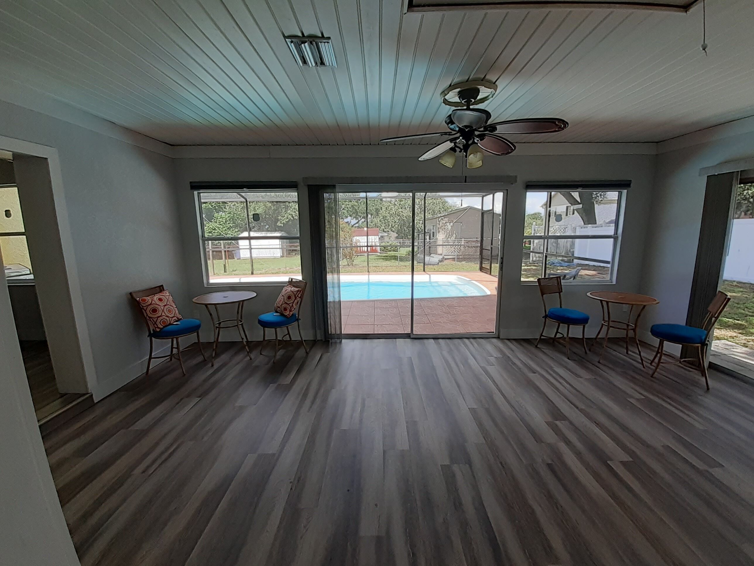 A spacious room with a wooden floor, large sliding glass doors leading to a backyard with a pool, and a ceiling fan.