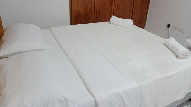 Una cama bien hecha con sábanas blancas, un armario de madera y una pared blanca.