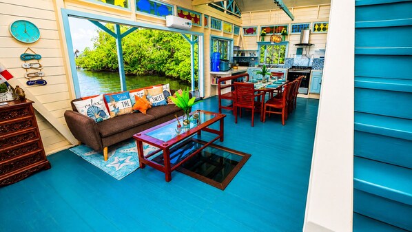 A room with a blue floor, a sofa with colorful cushions, a glass coffee table, and a view of greenery through large windows.
