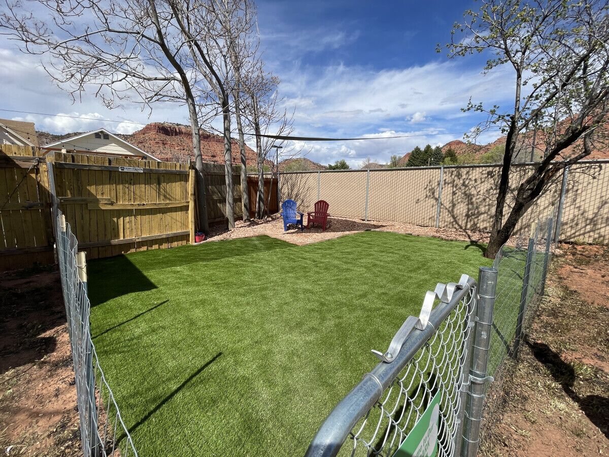 Un jardín trasero cercado con césped artificial, un cobertizo de madera y una silla roja.
