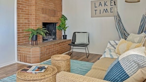 Ein gemütliches Wohnzimmer mit Kamin aus Backsteinen, KorboTTomanen, einer gestreiften Hängematte und einem Schild mit der Aufschrift 'LAKE TIME'.