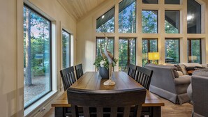 A dining area with a wooden table and chairs, large windows, and a sofa. - Campfire Dreams: Arcade, Hot Tub, Playset, 3BD/3Bath, Pet Friendly (Broken Bow)