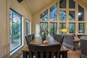 A dining area with a wooden table and chairs, large windows, and a sofa. - Campfire Dreams: Arcade, Hot Tub, Playset, 3BD/3Bath, Pet Friendly (Broken Bow)