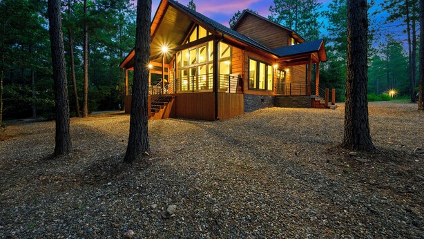 A wooden cabin with large windows, a covered porch, and a gravel driveway surrounded by trees. - Campfire Dreams: Arcade, Hot Tub, Playset, 3BD/3Bath, Pet Friendly (Broken Bow)