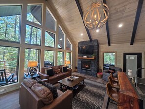 Un salon spacieux doté d’une cheminée, d’un canapé en cuir et de grandes fenêtres donnant sur une forêt.