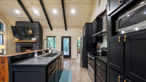 A modern kitchen with a stone fireplace, black cabinetry, and a large island. - Campfire Dreams: Arcade, Hot Tub, Playset, 3BD/3Bath, Pet Friendly (Broken Bow)