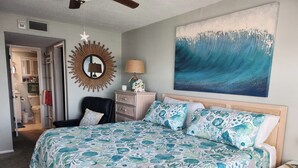 Un dormitorio con una cama grande, cabecera de madera, espejo de pared, mesita de noche y un cuadro de una ola.